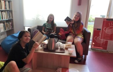 Trzy uśmiechnięte dziewczyny z książkami w ręku siedzą w bibliotece przy stoliku z herbatą
